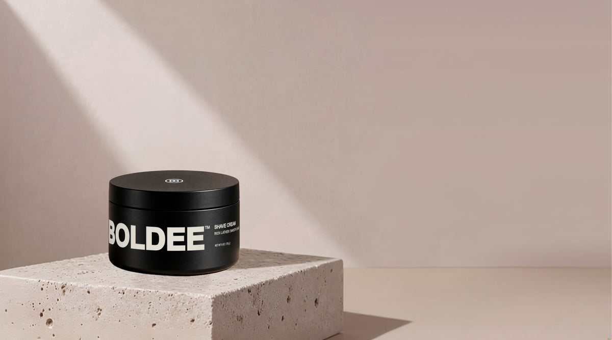 Matte black jar of BOLDEE Shave Cream displayed on a light stone pedestal against a beige background.